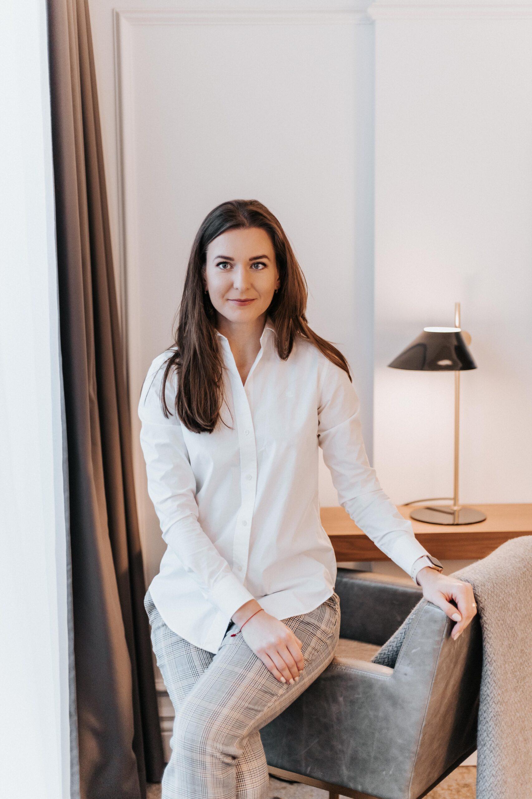 Portrait of interior designer Justina Kadžytė Garbštė dressed in white shirts and grey jeans, sitting on the armchair